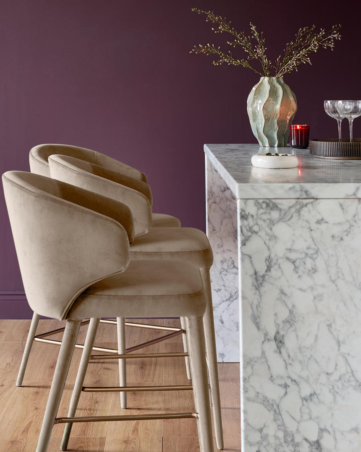 Champagne velvet bar stool with curved back and wooden legs at marble counter in styled interior setting.