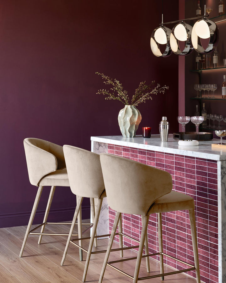 Champagne velvet bar stools with curved backs and wooden legs positioned at a modern marble and burgundy tile bar counter.