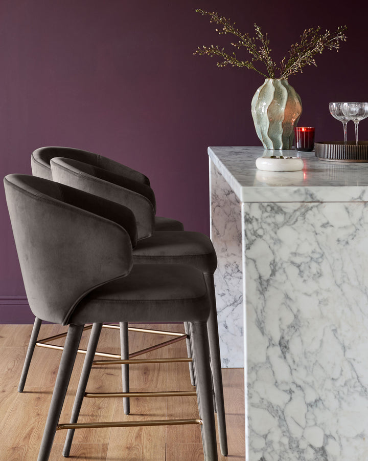 Ethan bar stool in dark chocolate velvet with curved back, metal legs, and footrest, shown at marble counter.