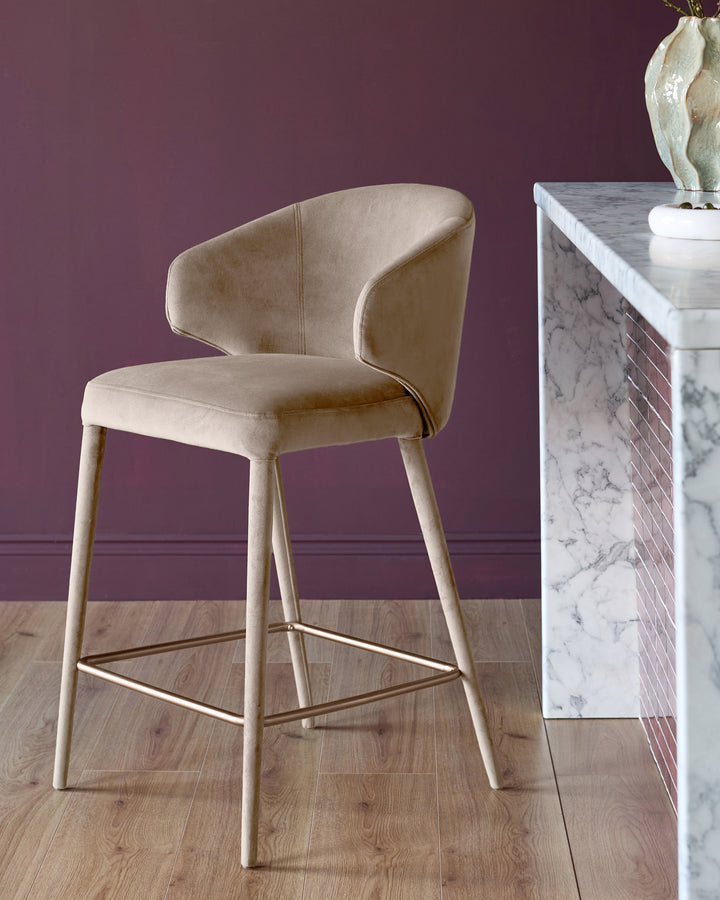 Champagne velvet bar stool with curved backrest, tapered wooden legs, and brass footrest against purple wall.