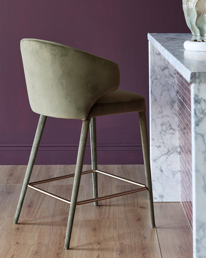 Ethan bar stool in olive green velvet with wooden legs and metal footrest, shown from rear angle against purple wall.