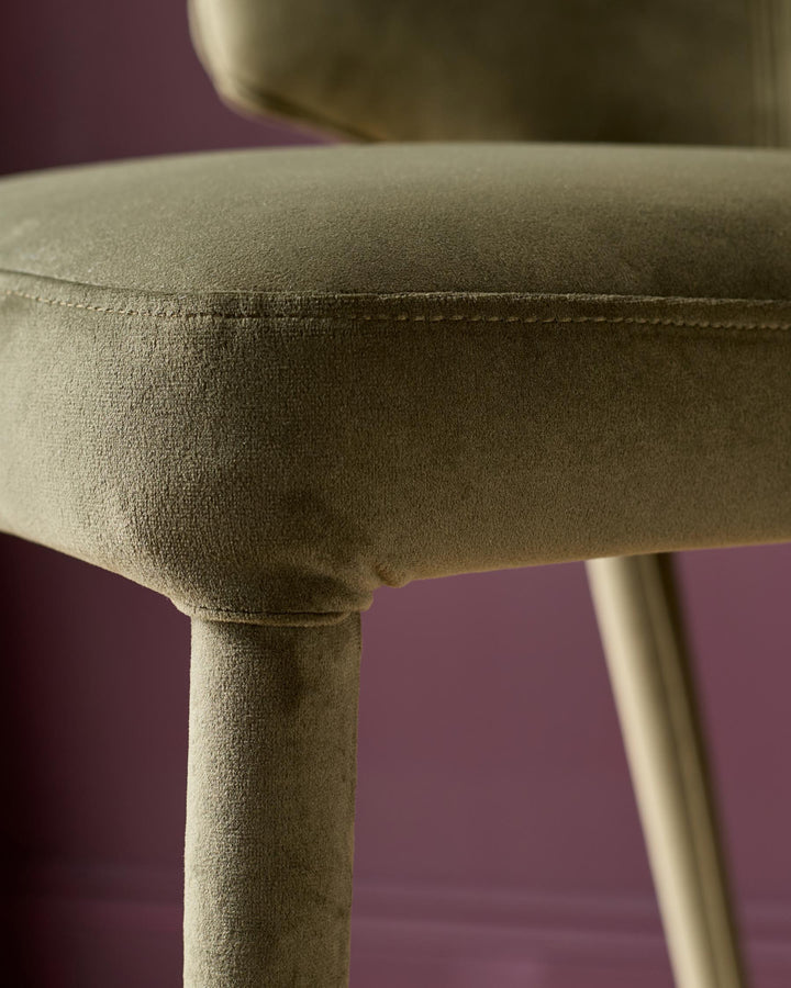 Ethan bar stool in olive green velvet, close-up detail of seat and wooden leg against purple background.
