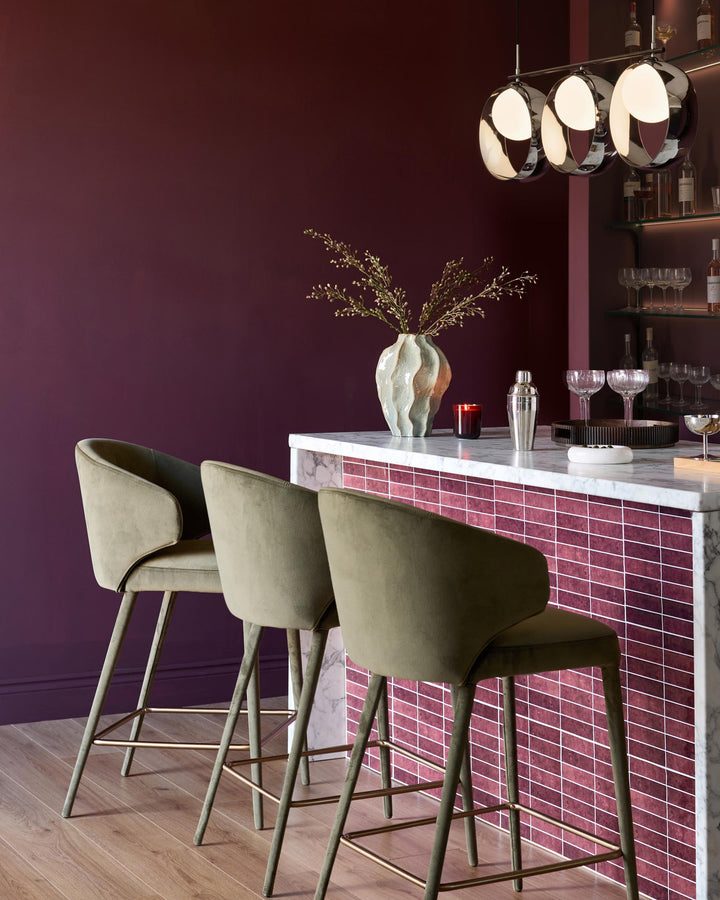 Ethan bar stool in olive green velvet with tapered legs, shown at burgundy tiled bar counter in modern interior setting.