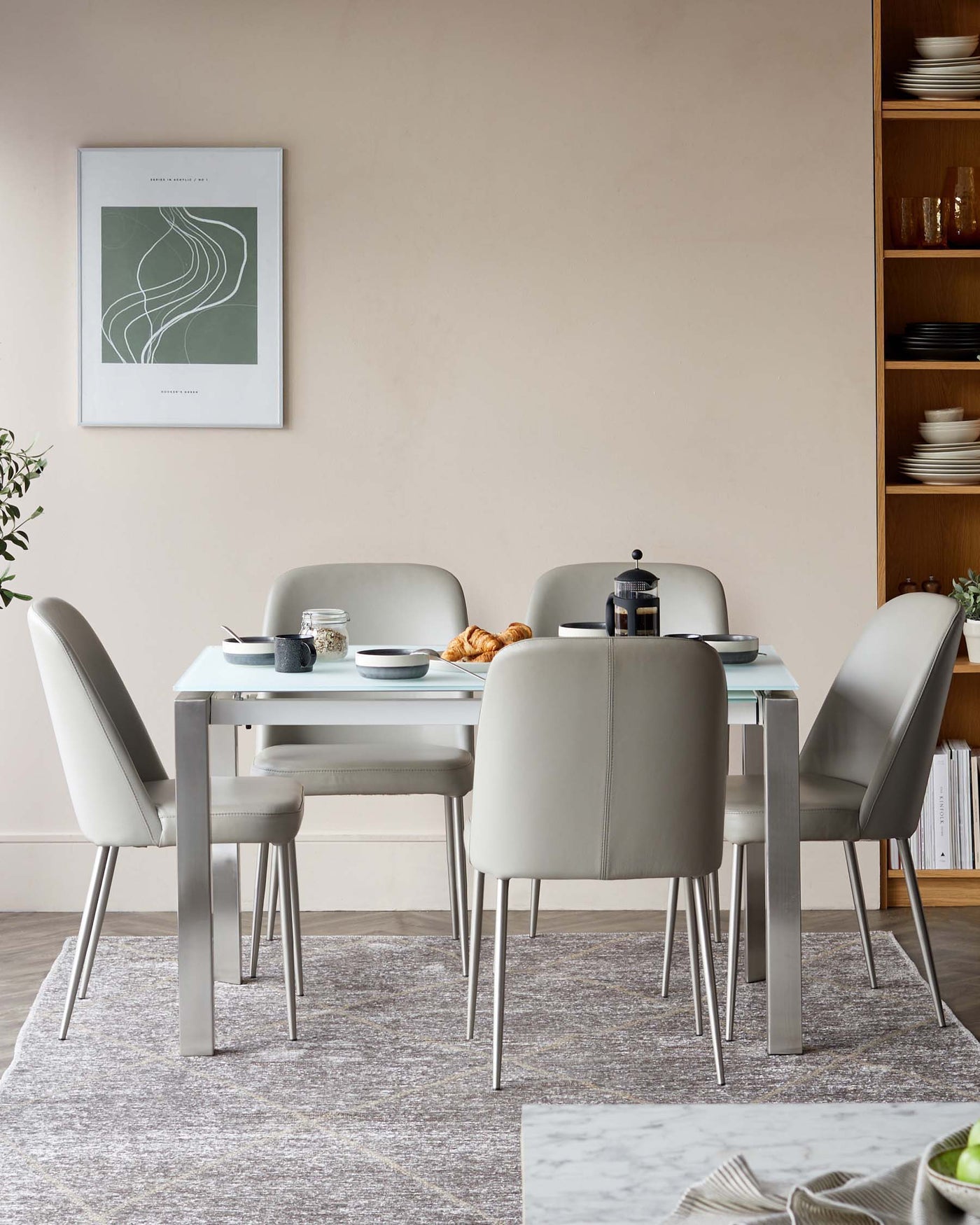 Modern minimalist dining set with a rectangular light grey tabletop and sleek stainless steel legs, surrounded by four upholstered chairs in matching tone with curved backs and slim metallic legs, arranged on a textured grey area rug. A wooden open shelf cabinet with various dishes is partially visible in the background.