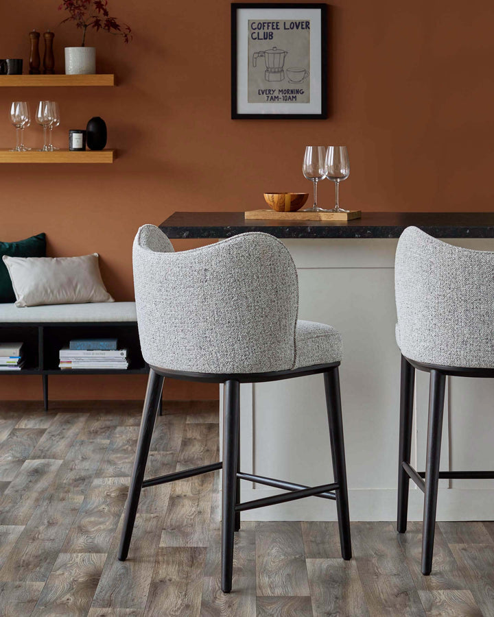 Two grey upholstered bar stools with black legs sit in front of a breakfast bar with decorative shelves and seating nearby.