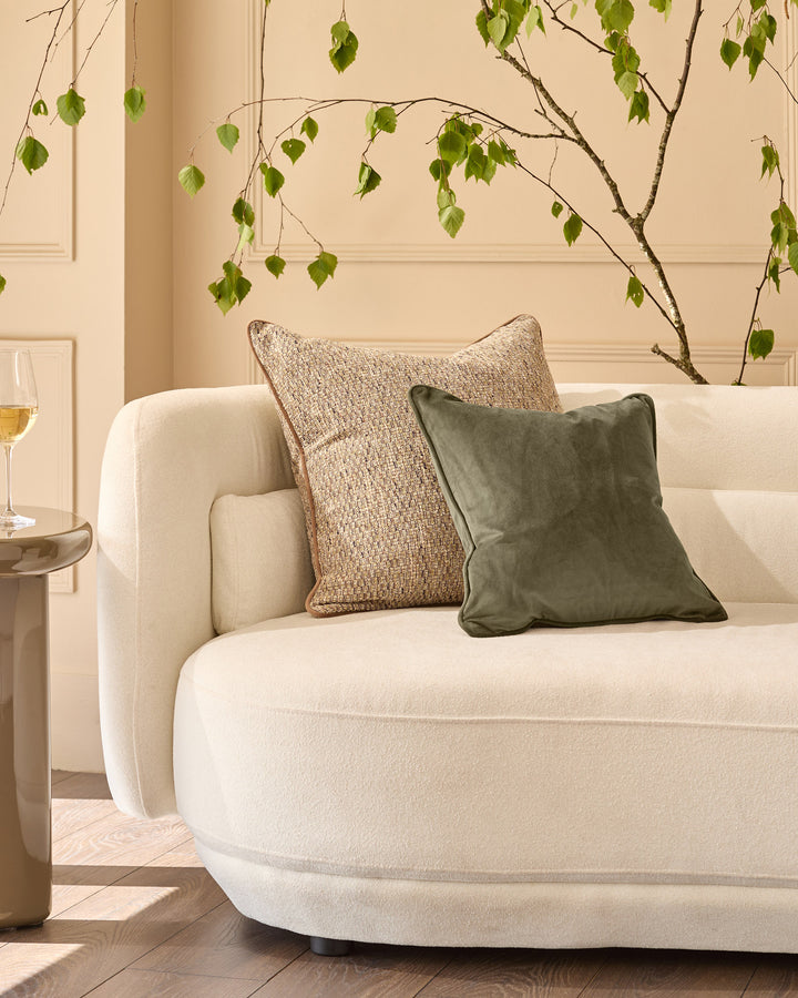 Large green velvet cushion displayed on a cream curved sofa with natural styling and hanging foliage decoration.