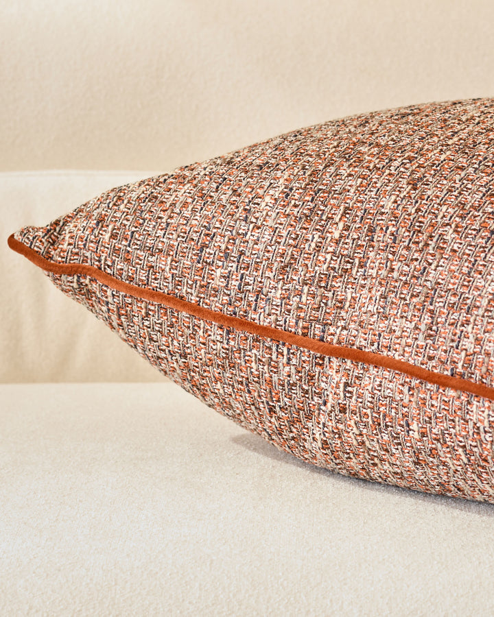 Rust-colored woven lumbar cushion with cream trim, displayed at an angle against a neutral background.