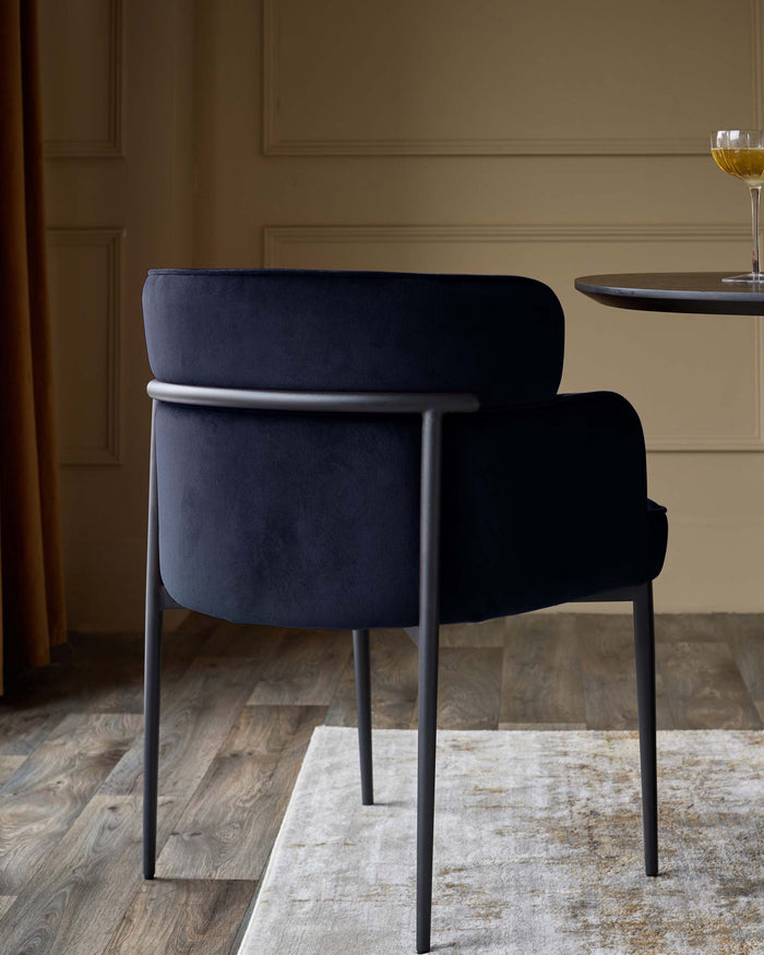 A navy velvet chair with a matte black frame, placed partially on a patterned rug near a round table and a glass of wine.