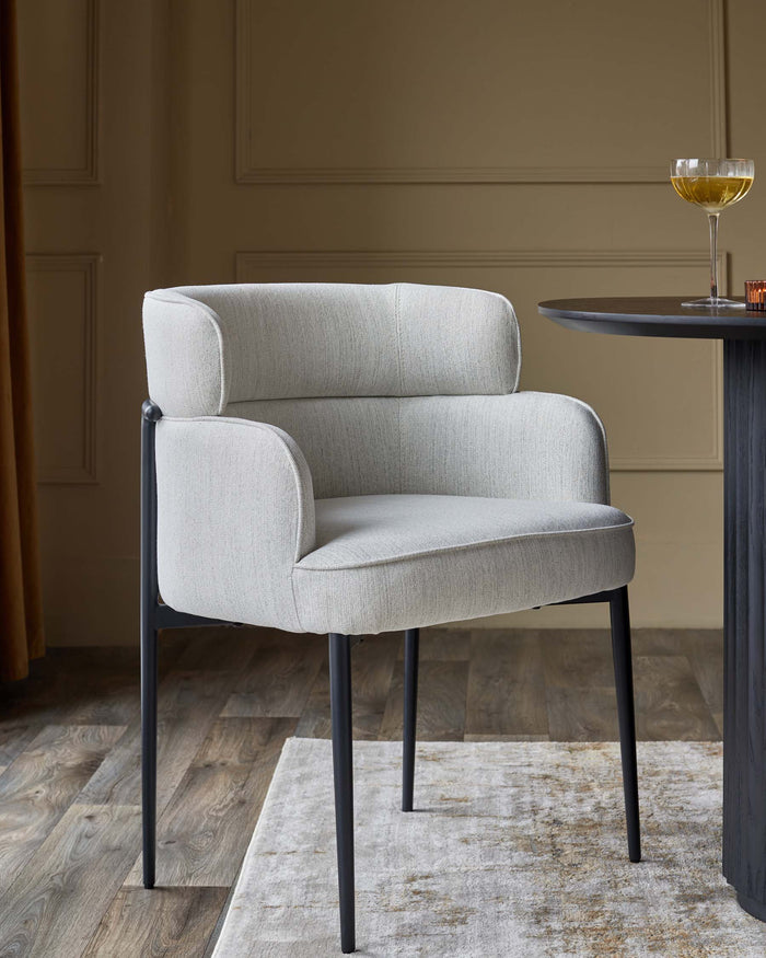 A stylish, light grey upholstered armchair with a modern black frame, positioned beside a dark wood table with a drink.