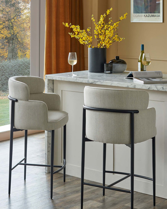 Two elegant bar stools with oatmeal fabric and black metal frames sit at a marble-topped island in a light, airy kitchen.