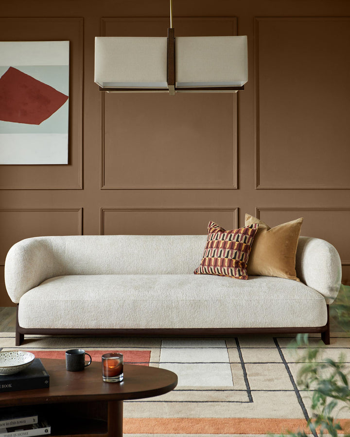 A modern, light-coloured sofa with two accent cushions. Features a brown wooden frame and a contemporary rug.
