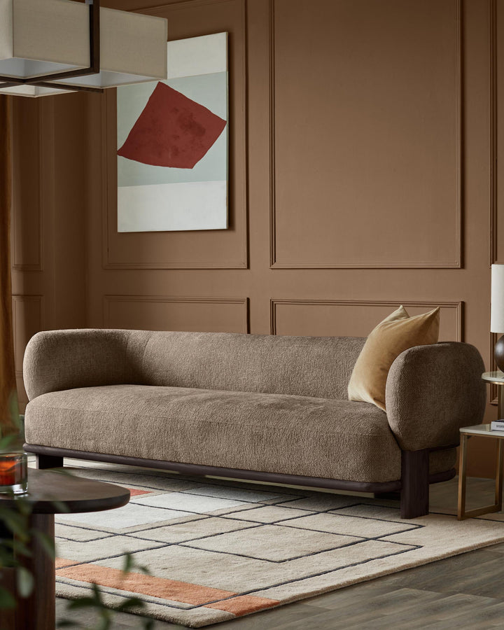A stylish, brown sofa with a neutral pillow sits on a patterned rug in a room with brown walls and modern art.
