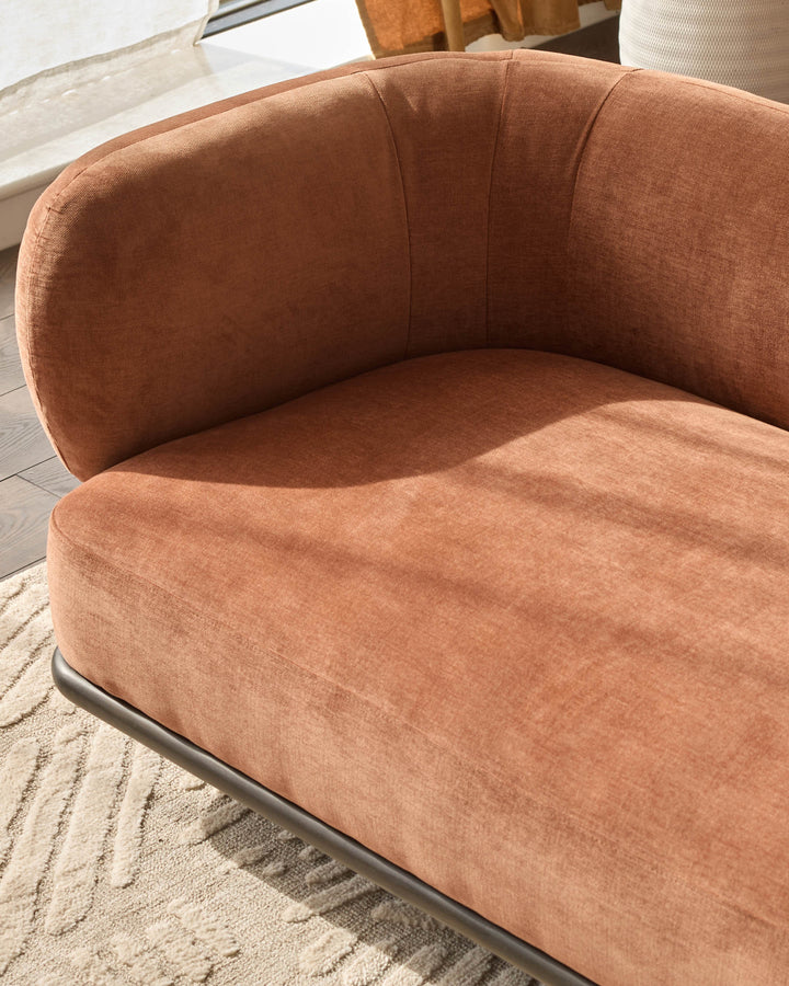 Apricot upholstered curved armchair with rounded backrest and seat, shown at an angle on a neutral rug in bright natural light.