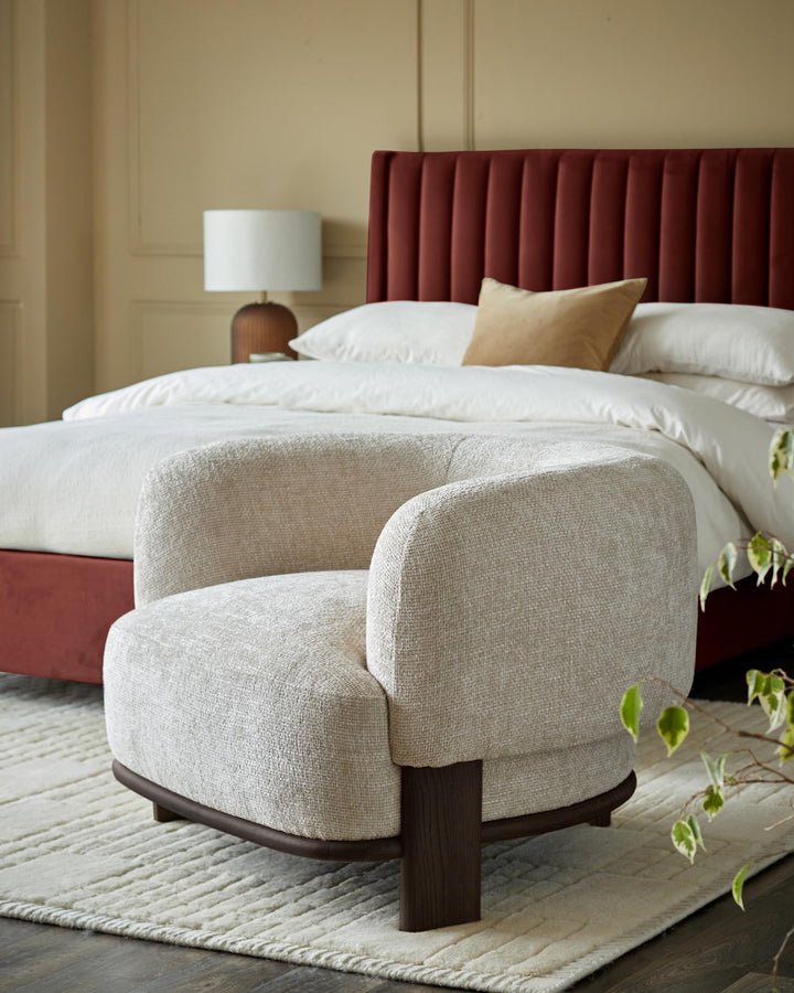 A stylish bedroom featuring a red velvet bed with a neutral-toned chair and patterned rug.
