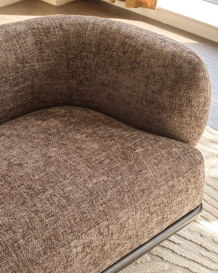 Mocha fabric curved armchair with rounded backrest and seat, metal base, angled side view on light wood flooring.