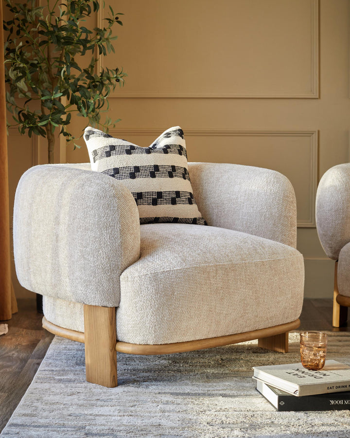 A cosy armchair with a textured fabric, complemented by a patterned cushion and wooden frame. Books and a drink sit nearby.