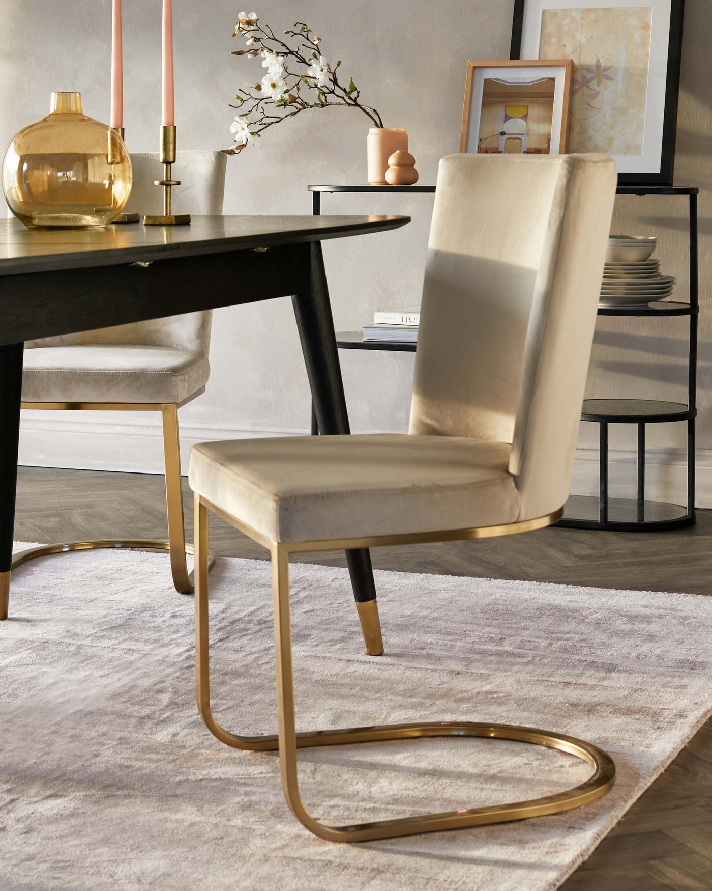 Elegant contemporary dining room featuring a sleek black oval-shaped dining table with a matte finish, complemented by luxury upholstered dining chairs in a soft beige tone with unique gold-finished metal cantilever bases. A side table with shelves displays decorative items, enhancing the modern aesthetic of the space.