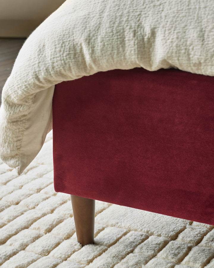 Burgundy velvet ottoman with cream knit throw blanket, wooden leg, on textured cream rug.