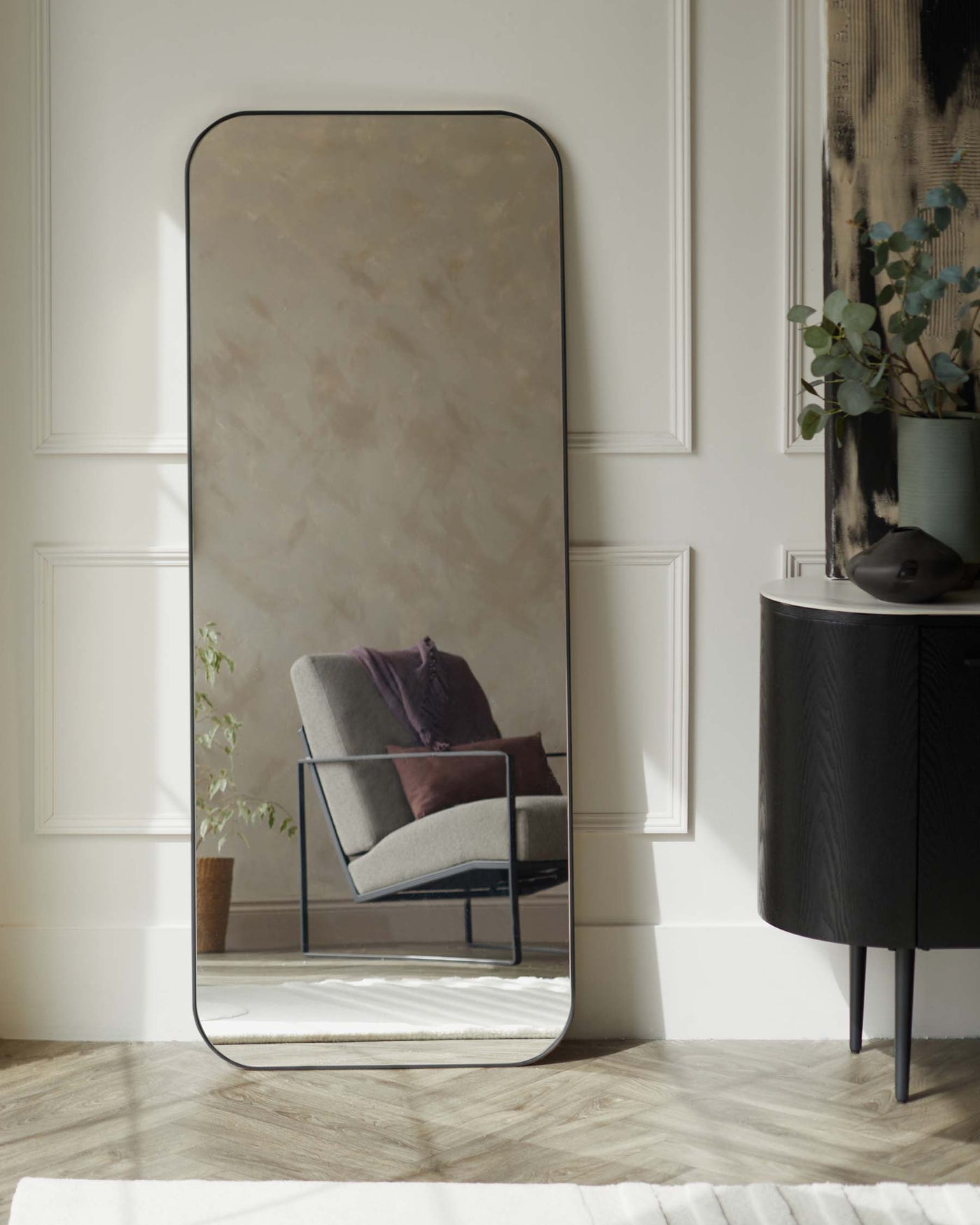 A large, rectangular floor mirror with rounded top corners and a thin black frame standing against a wall, reflecting a modern armchair with a charcoal upholstered seat and a minimalist black metal frame. Beside the mirror is a small, round, black side cabinet with slender legs and textural front panels.