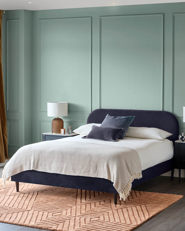 A bedroom with a plush, dark blue bed, white linen, side tables, and a patterned rug. A calming and stylish space.
