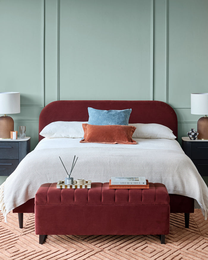 Bedroom with a deep red bed and ottoman, bedside tables, lamps, and a patterned rug, creating a cosy, stylish interior.