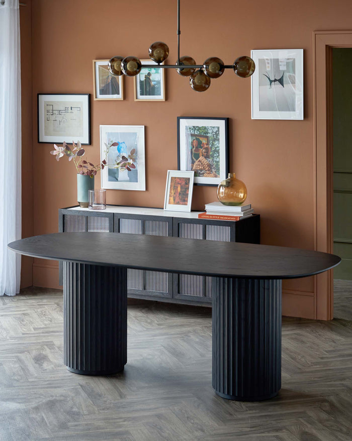 A sleek oval dining table with fluted columns, paired with a textured sideboard, surrounded by framed artwork and a modern light fixture.