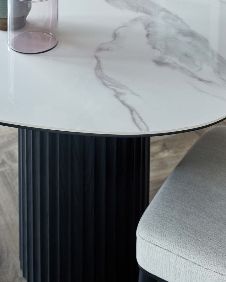 Round dining table with a white marble top featuring subtle gray veining, supported by a ribbed black base, alongside a plush gray chair.
