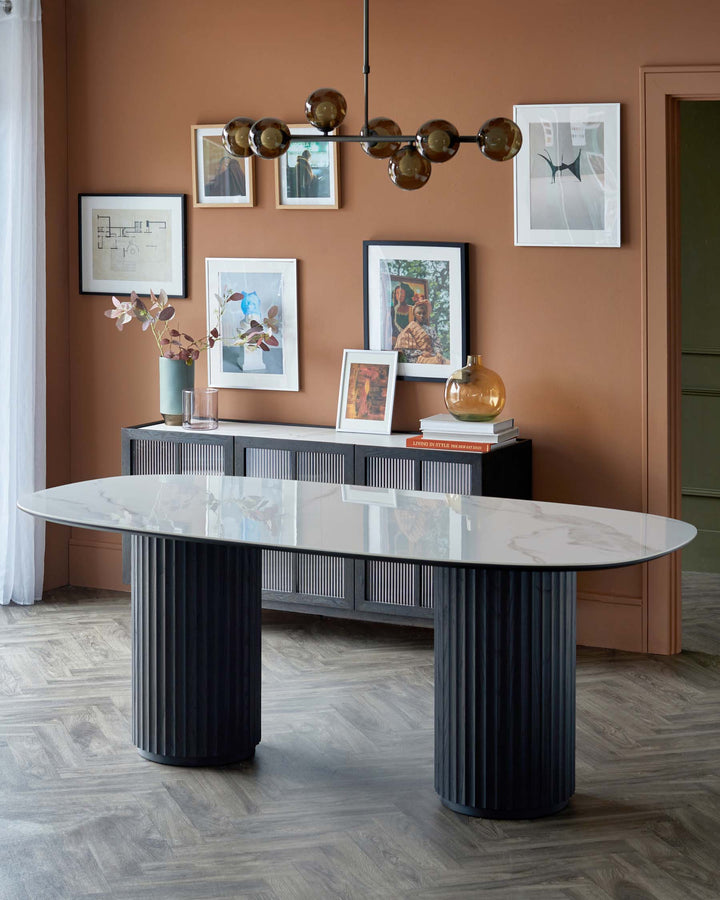 A sleek oval dining table with a marble top and ribbed black bases, paired with a modern sideboard and artistic wall decor.