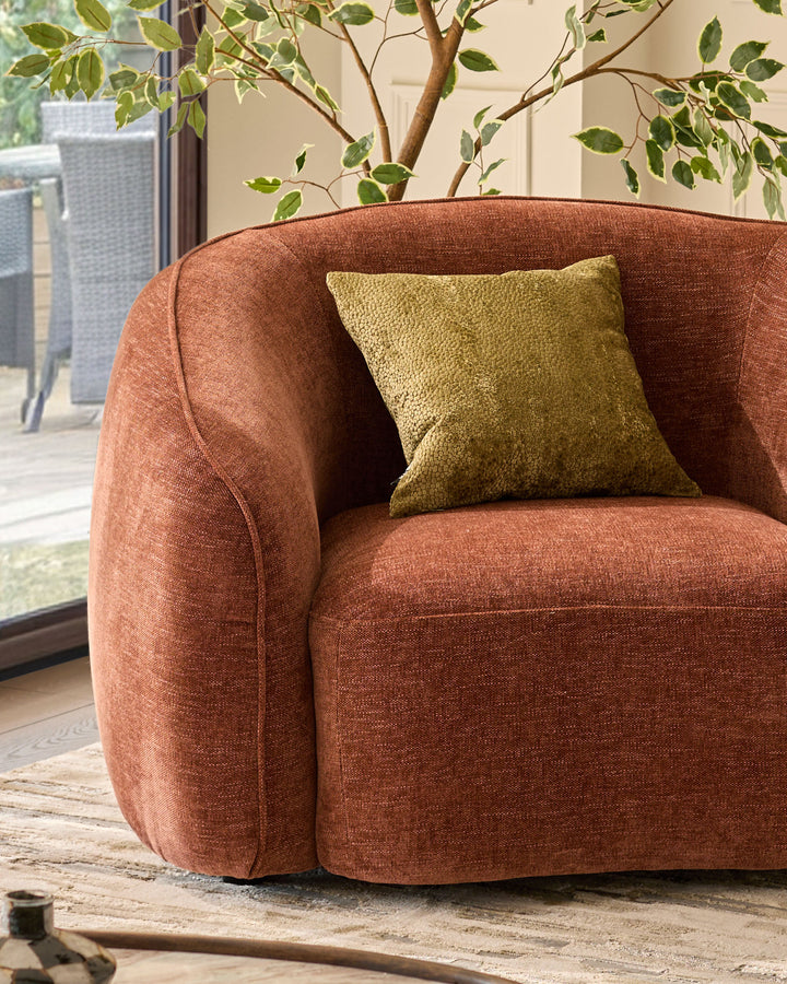 Harlene cushion in medium olive green, displayed on a rust-colored curved armchair in a bright interior space.