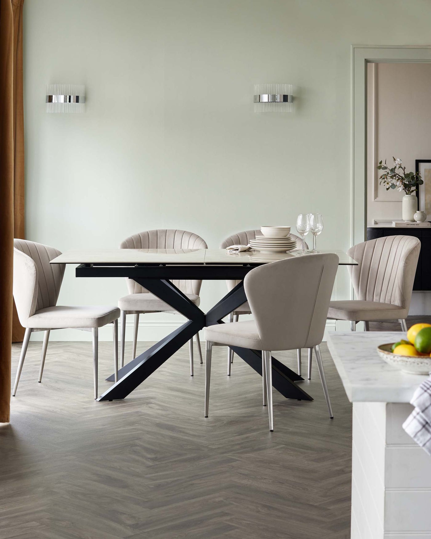 A modern dining set with a rectangular table featuring a black top and a bold X-shaped metal base. Includes six contemporary upholstered chairs in a soft beige tone, with a comfortably curved backrest and slender metal legs. The set is displayed in an elegantly simple room with a light grey wood flooring.