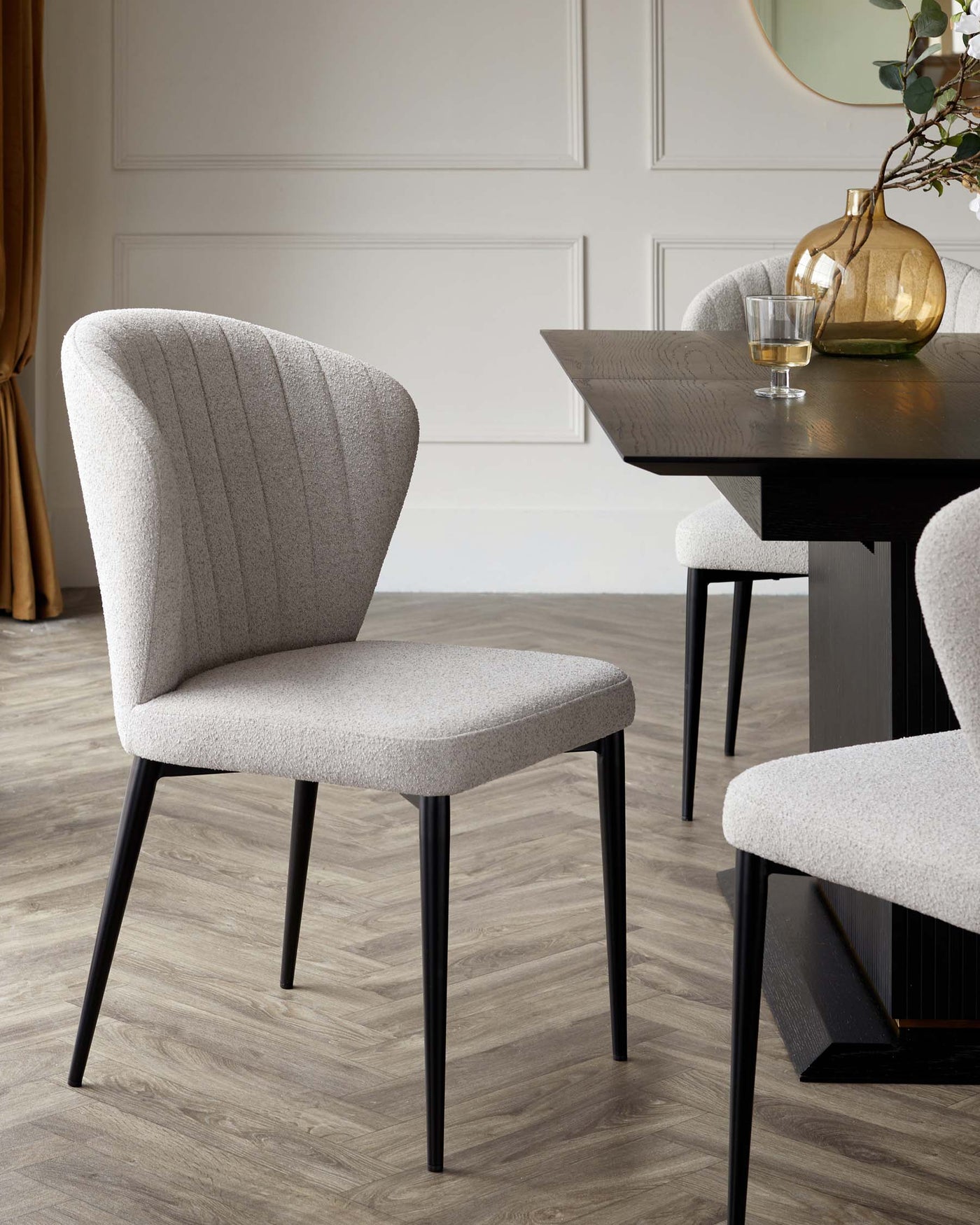 Elegant dining chair with textured light grey upholstery and a curved, high-back design on tapered black legs. Dark wooden dining table with prominent wood grain texture visible, hosting a decorative vase and a glass of a drink. The setting suggests a sleek, modern aesthetic.