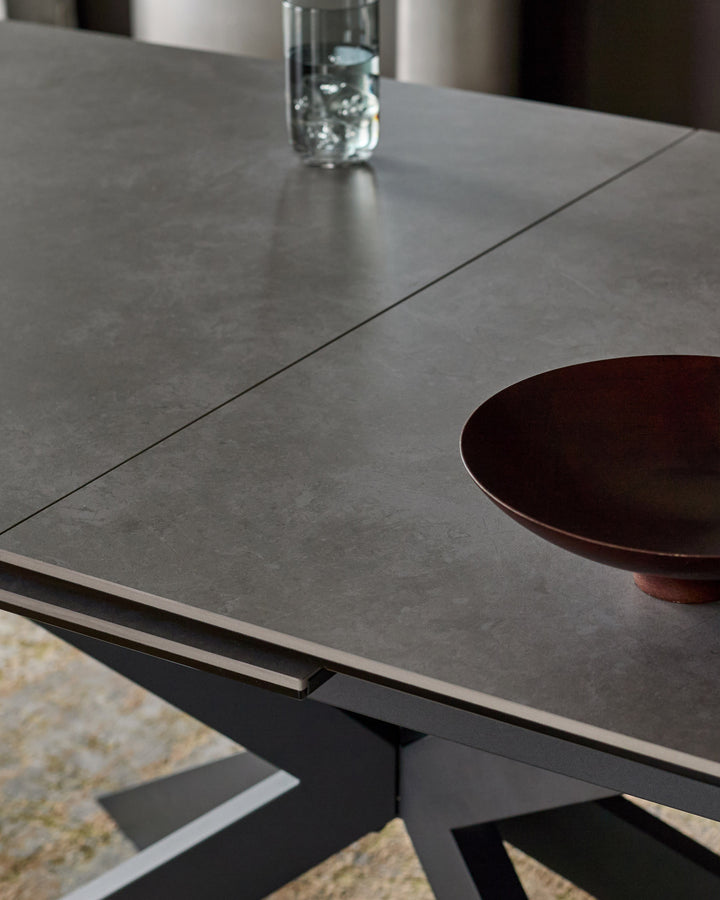 Hayden extending dining table in light grey ceramic with black base, shown at angle with place setting and water glass.
