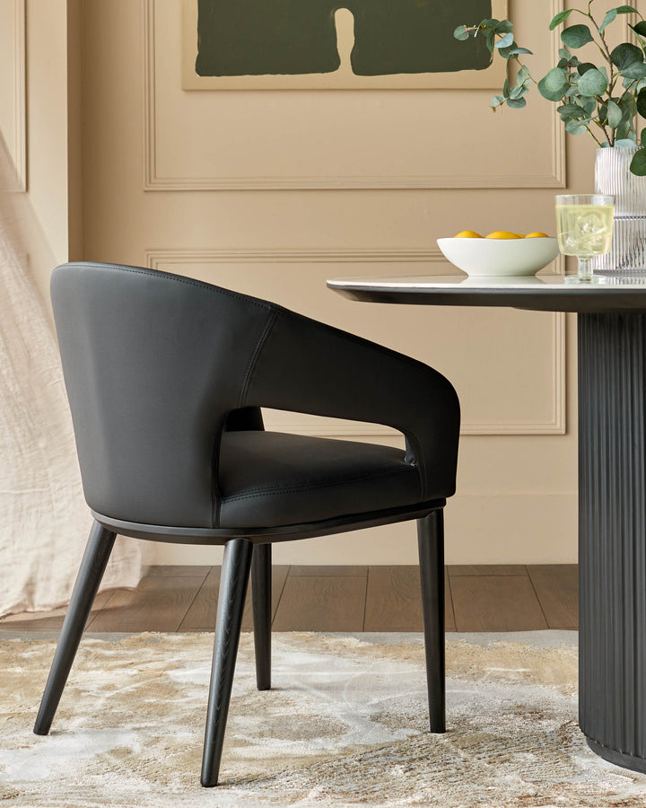 A modern black leather chair sits beside a round table with fruit and a vase of eucalyptus, set on a cream rug.
