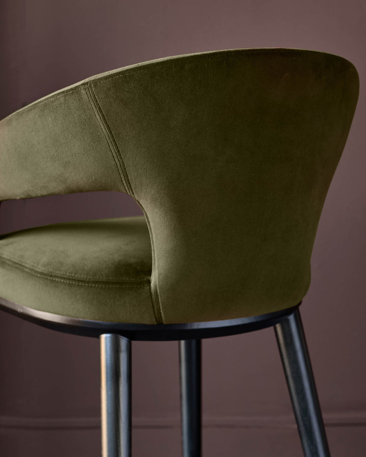 An upholstered bar stool with a curved back in a moss green velvet, featuring a metal base and legs.