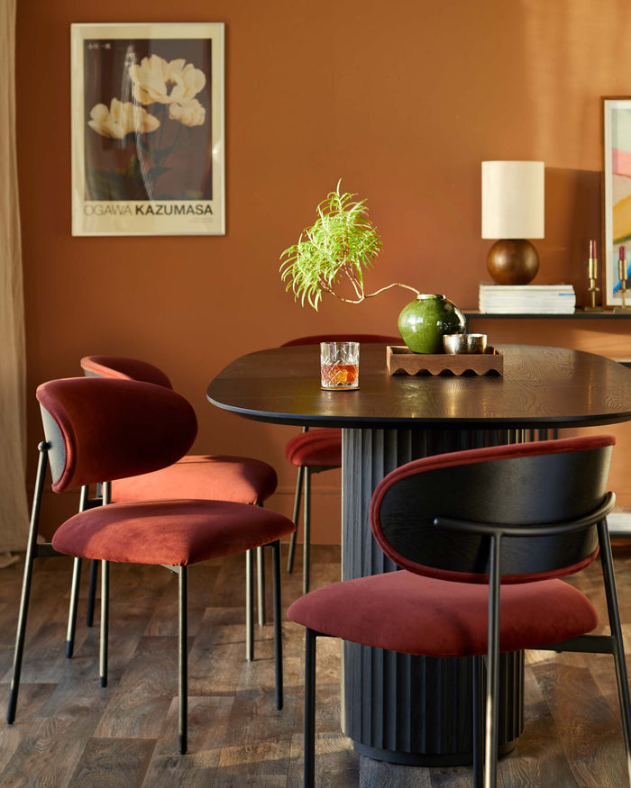 A modern dining set featuring a round, dark wood table and four stylish chairs with maroon velvet seats and black frames.