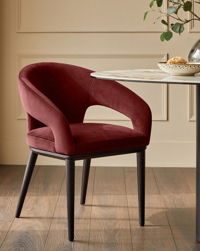 A stylish, contemporary dining chair in deep red velvet, with dark wooden legs, sits beside a round marble-topped table.