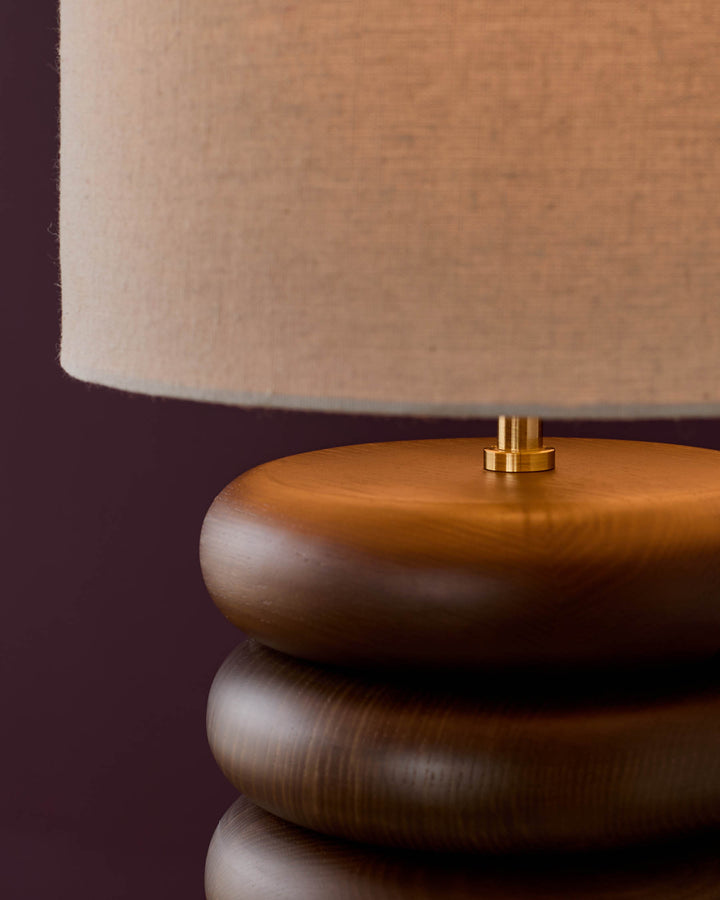 Hepworth table light with stacked dark wood base, brass connector, and natural linen shade against burgundy background.