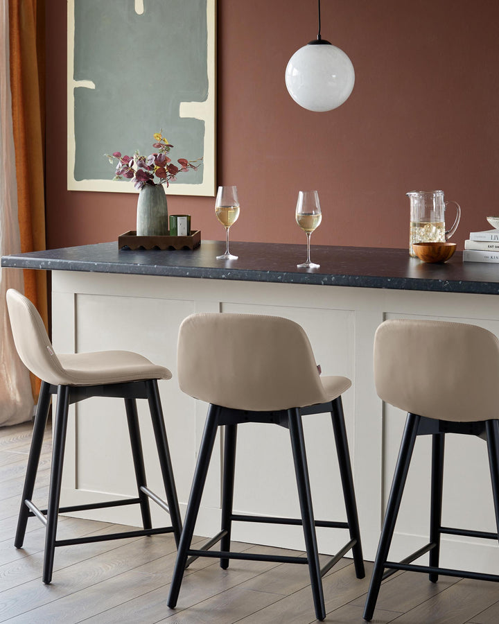 Three counter stools with black legs sit beneath a black countertop and cream island. Brown wall and light fixture behind.