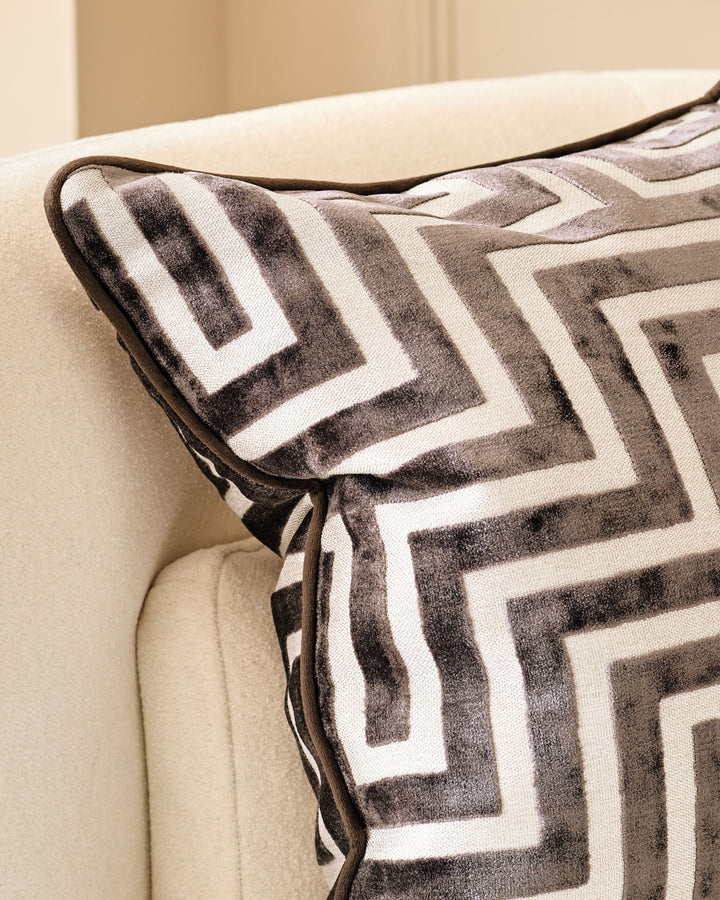 Imani cushion in chocolate with geometric striped pattern, displayed on cream upholstered furniture with leather piping detail.