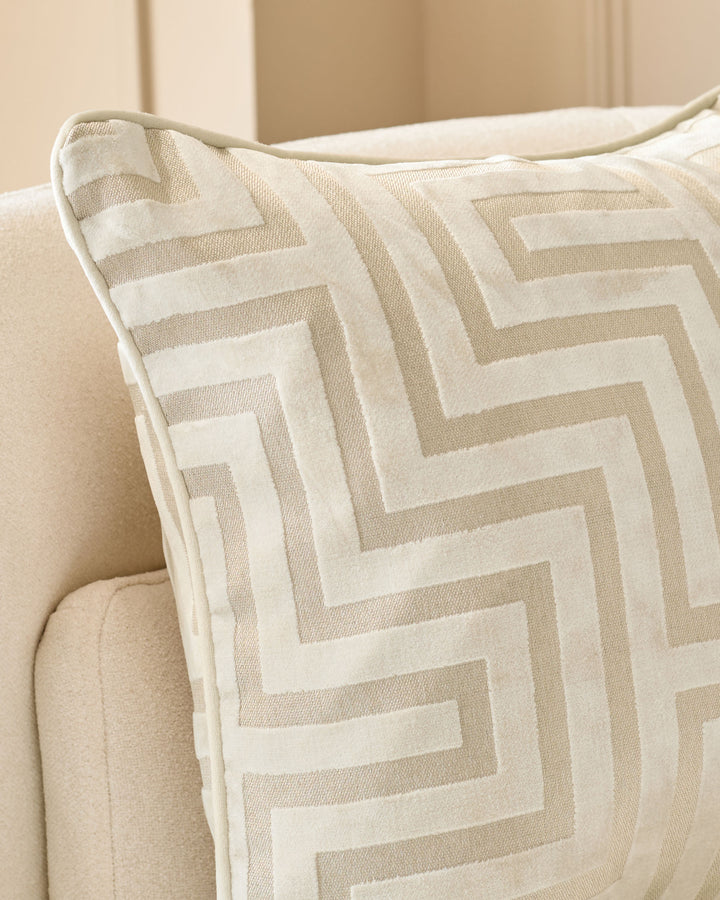 Imani medium ivory cushion with geometric striped pattern, displayed on a beige upholstered chair.