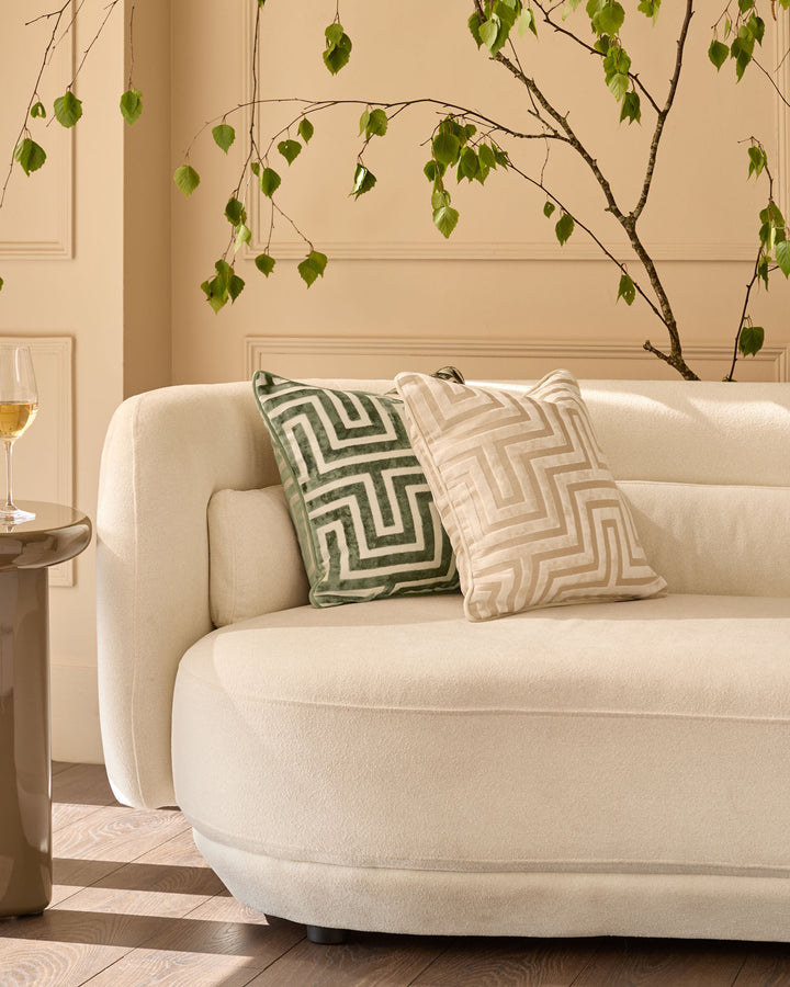 Imani cushion in olive green with geometric pattern, displayed on cream curved sofa in modern living room setting.
