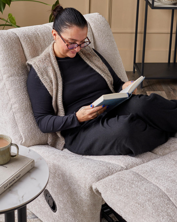 A woman relaxing on a light beige recliner armchair. A side table with a book and mug are visible.
