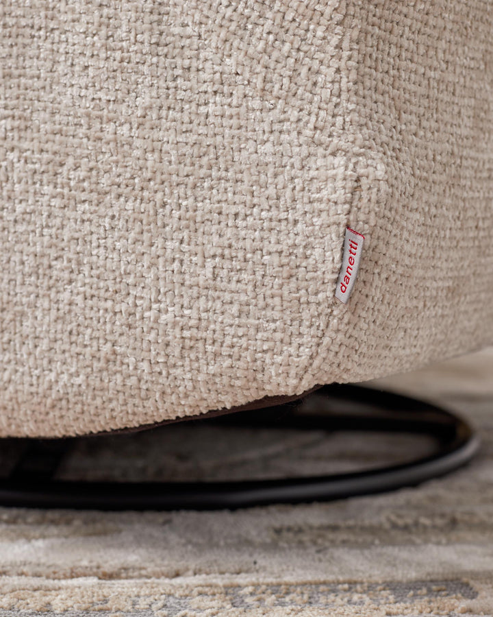 Close-up of a textured, beige upholstered chair with a 'danetti' tag, and a glimpse of the swivel base and patterned rug beneath.