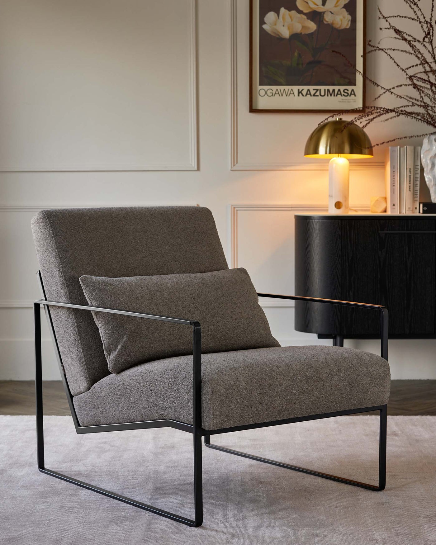 Modern minimalist armchair with a sleek black metal frame and comfortable heathered grey upholstery, positioned in a stylish interior setting beside a black cabinet with a brass-domed lamp and framed artwork on the wall.