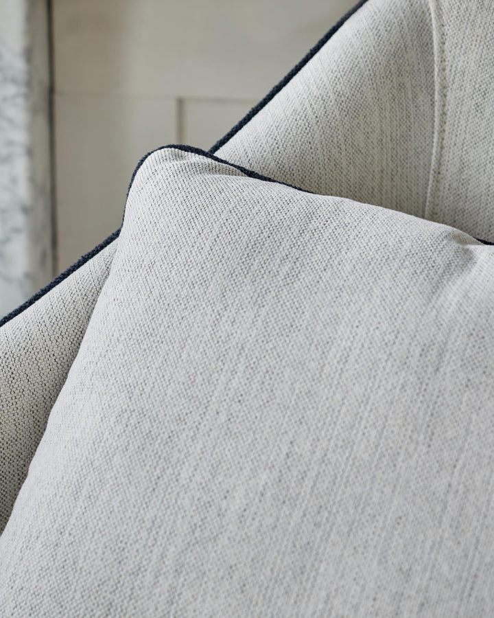 Close-up of a textured, light grey cushion with dark piping detail, resting against a larger upholstered furniture piece.