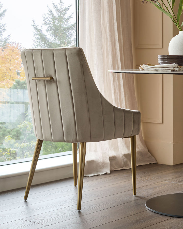 Champagne velvet dining chair with vertical channel tufting, brass legs, and gold handle detail, shown in modern room setting.