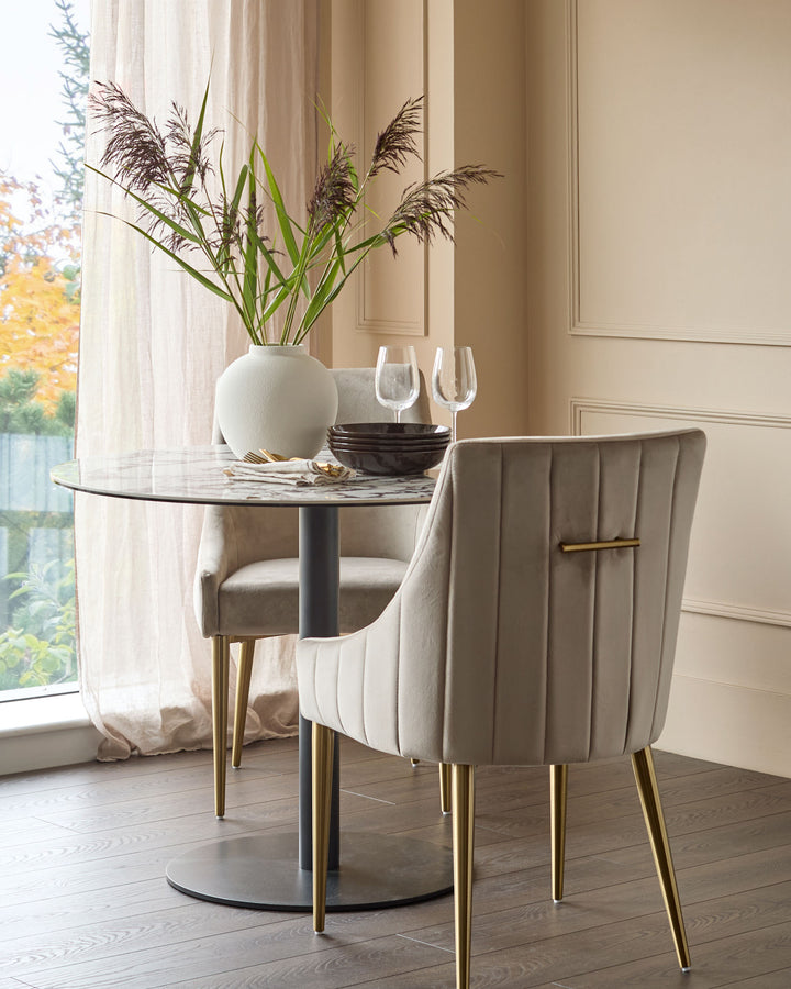Champagne velvet dining chair with brass legs, shown at round marble table with white vase and glassware in bright room.