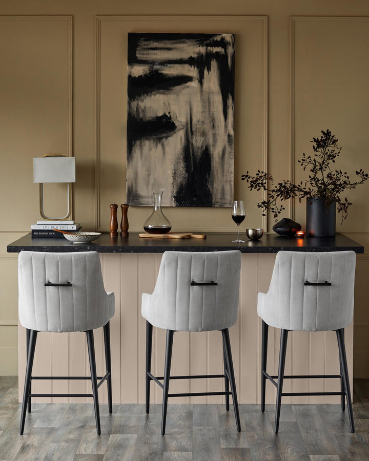 Three grey bar stools with black legs sit at a kitchen island with a black worktop and artwork.