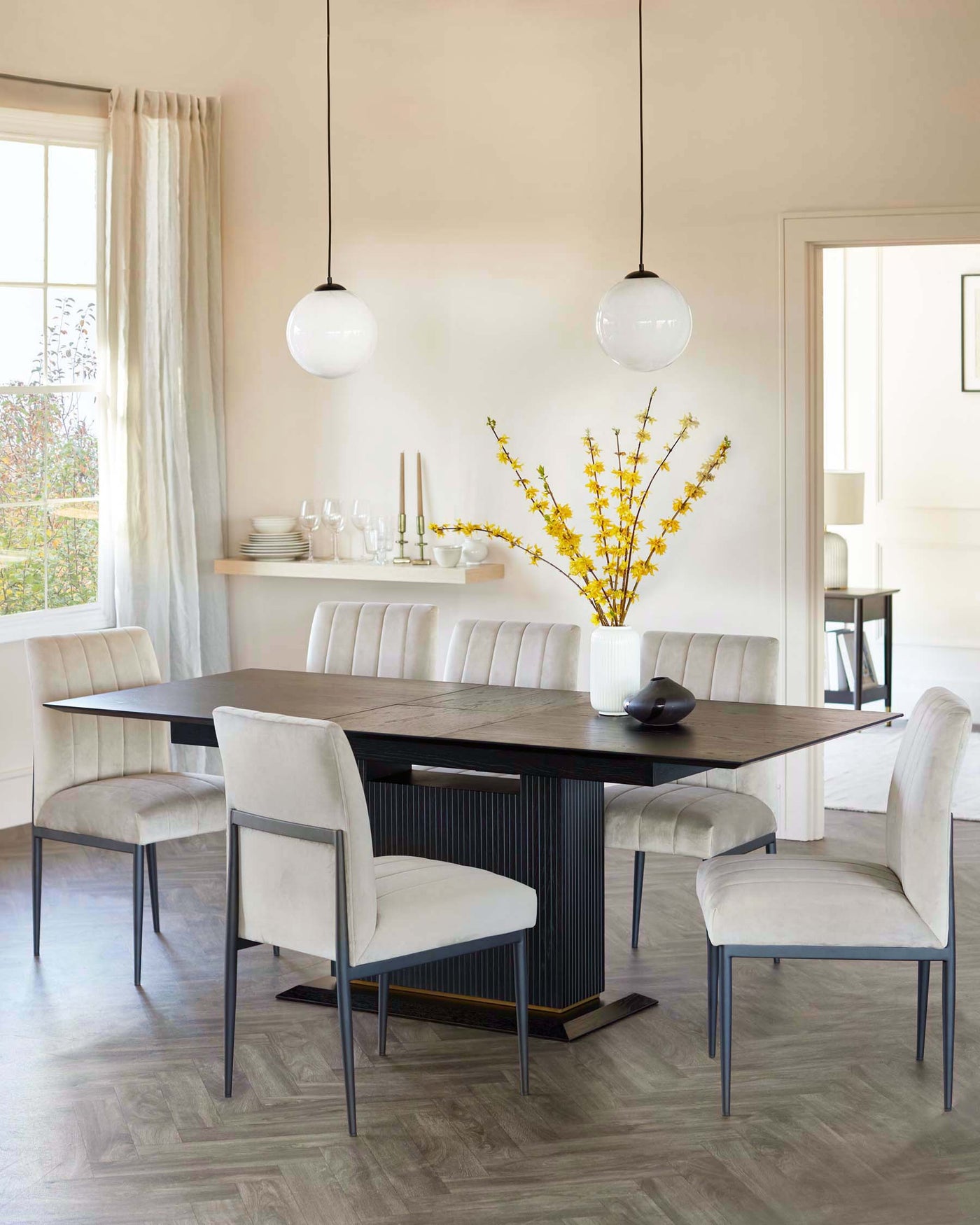 A modern dining room showcasing a dark wood rectangular dining table with a fluted design on the sides and an elegant metal base. Six upholstered dining chairs with soft fabric and slim metal legs are positioned around the table. The room is complemented by minimalist decor and two simple, round pendant lights overhead.