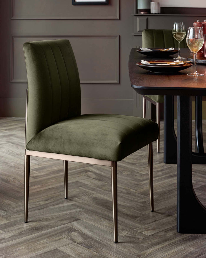 A green velvet dining chair with brushed metal legs stands by a dark wood dining table laid for a meal.